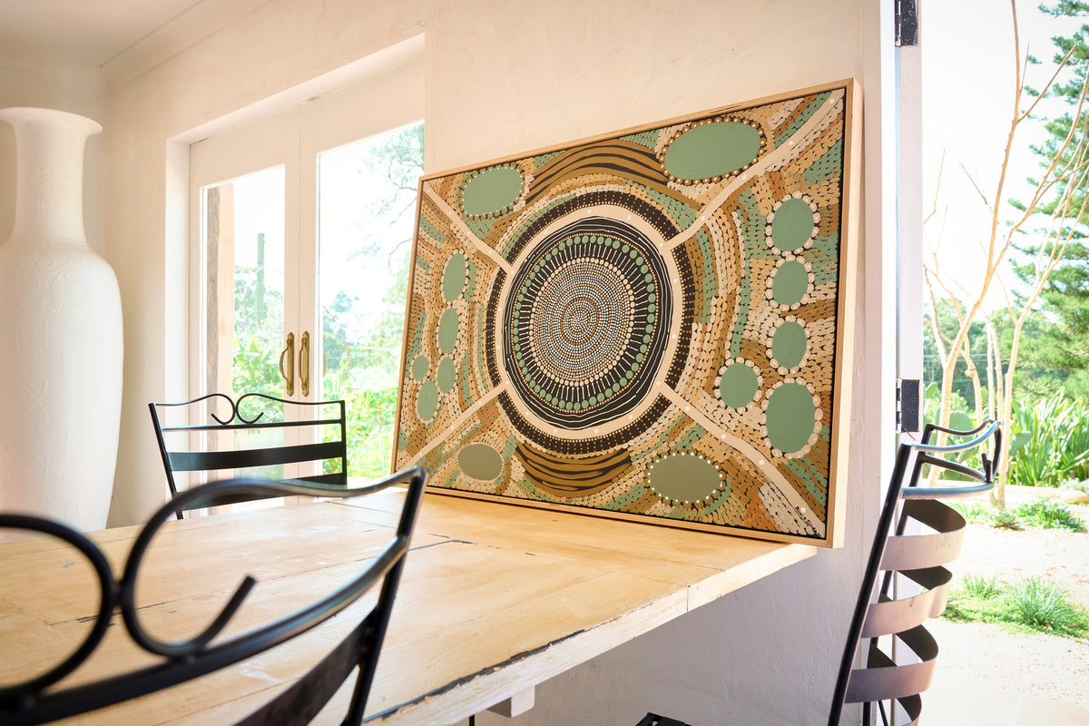 A large framed square wall art piece with an intricate circular mandala-style pattern in green, beige, brown, and black, leaning against a wall in a dining room next to a wooden table and chairs.