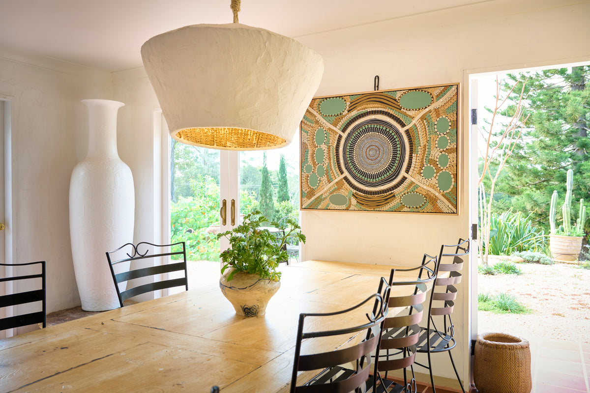 A large square wall art piece with a green, beige, and brown mandala-style geometric pattern hangs on a cream wall in a bright dining room. The room features a wooden dining table, black metal chairs, a large white ceramic vase, and a textured cream pendant light hanging above.