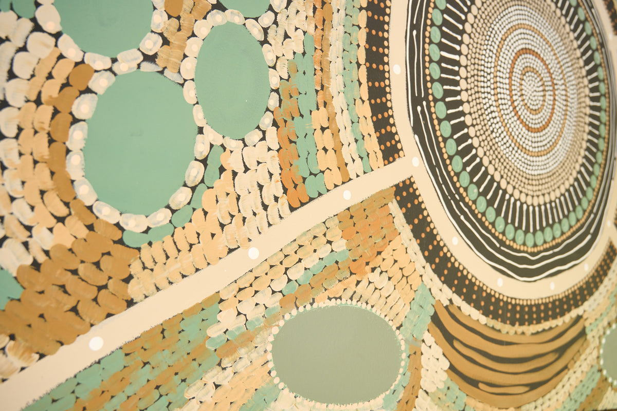 A close-up of a painted circular Aboriginal-style artwork with concentric dots, bands, and oval shapes in beige, brown, black, mint green, and white.
