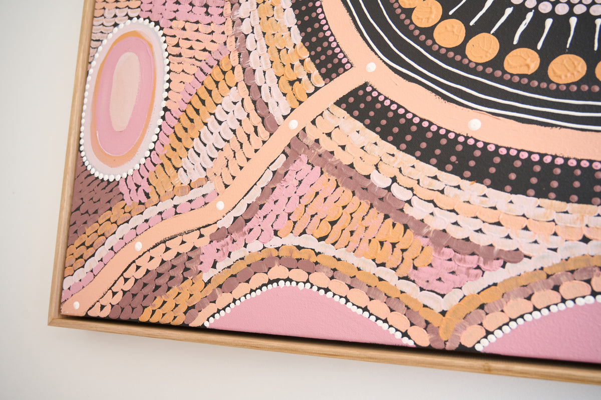 A close-up of a framed abstract artwork featuring pink, peach, beige, black, and white painted patterns on a canvas, mounted on a light wall.
