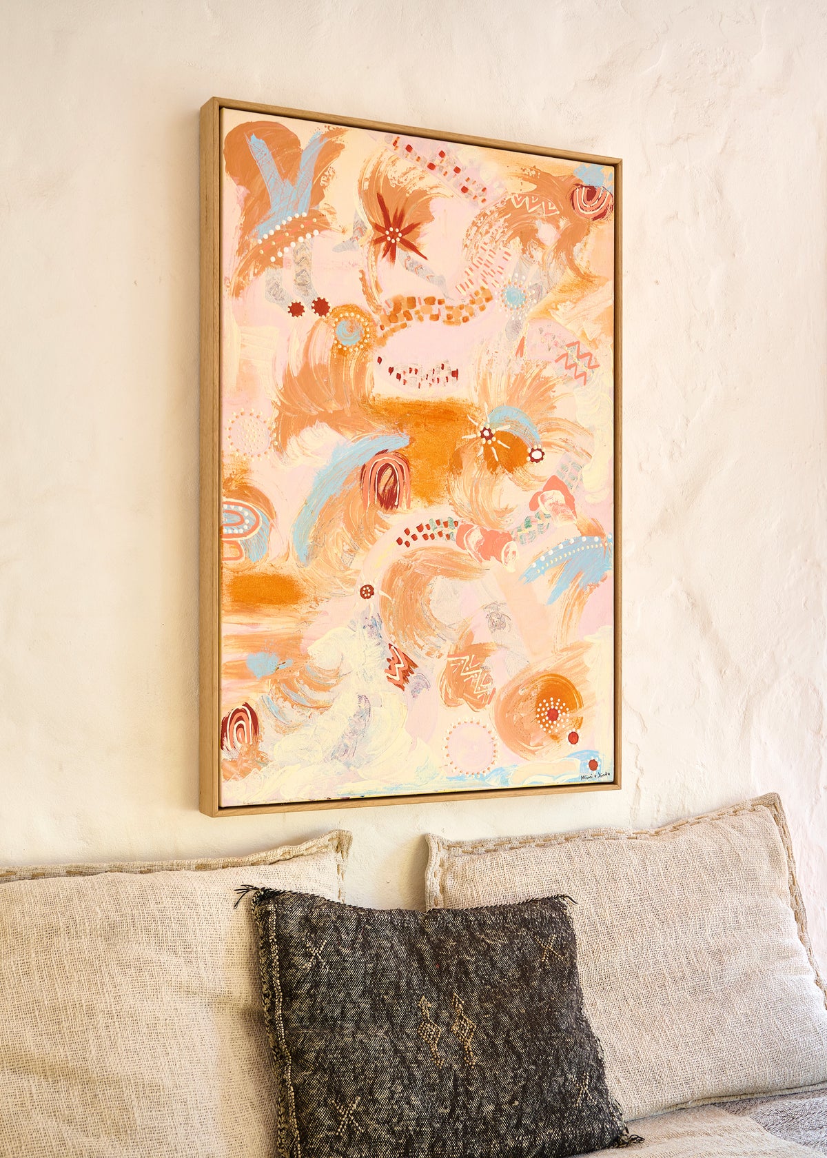 A framed abstract art print featuring warm orange, peach, and light blue brushstrokes on a cream background, hanging on a textured off-white wall above a sofa with neutral-toned cushions.