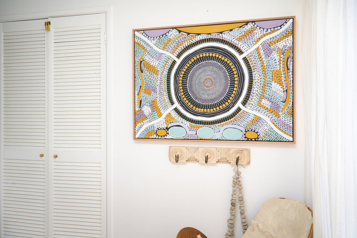 A large framed Aboriginal art print featuring a complex circular mandala design in yellow, black, white, and purple, hanging on a white wall above a wooden coat rack.