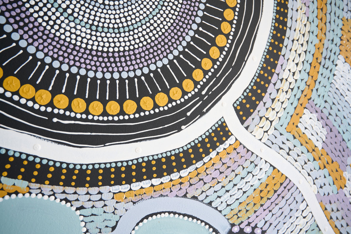 A close-up of an Aboriginal-style dot painting artwork featuring concentric circles, geometric patterns, and stylized figures in black, white, yellow, purple, and light blue.