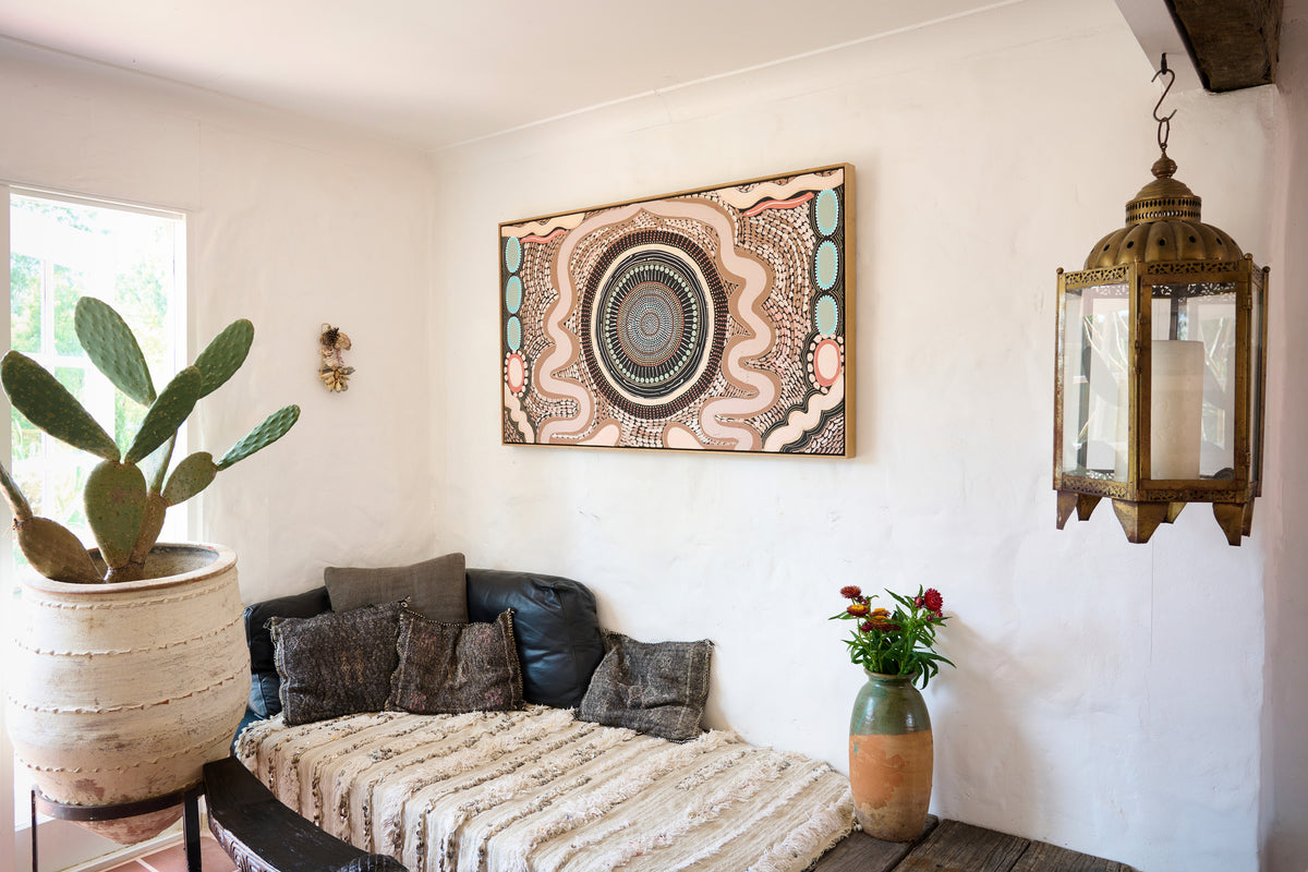 A framed bohemian-style circular artwork hangs on a white plaster wall above a dark leather daybed with textured beige throw pillows and a woven blanket. The room features a large potted prickly pear cactus on the left, a hanging brass lantern on the right, and a small vase of red flowers on the daybed.
