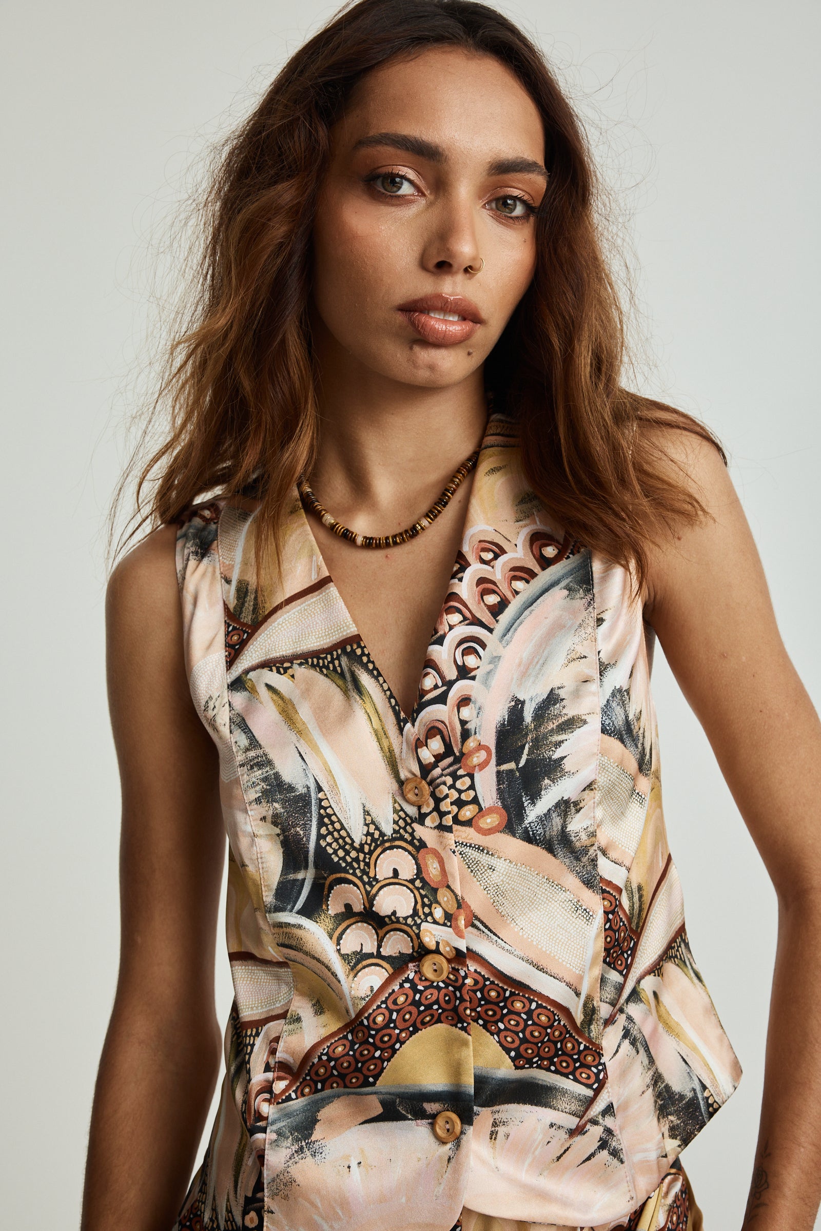 A female model wearing a sleeveless silk vest with a geometric and floral print, in tones of beige, white, and gold, with a collar and buttonless front.