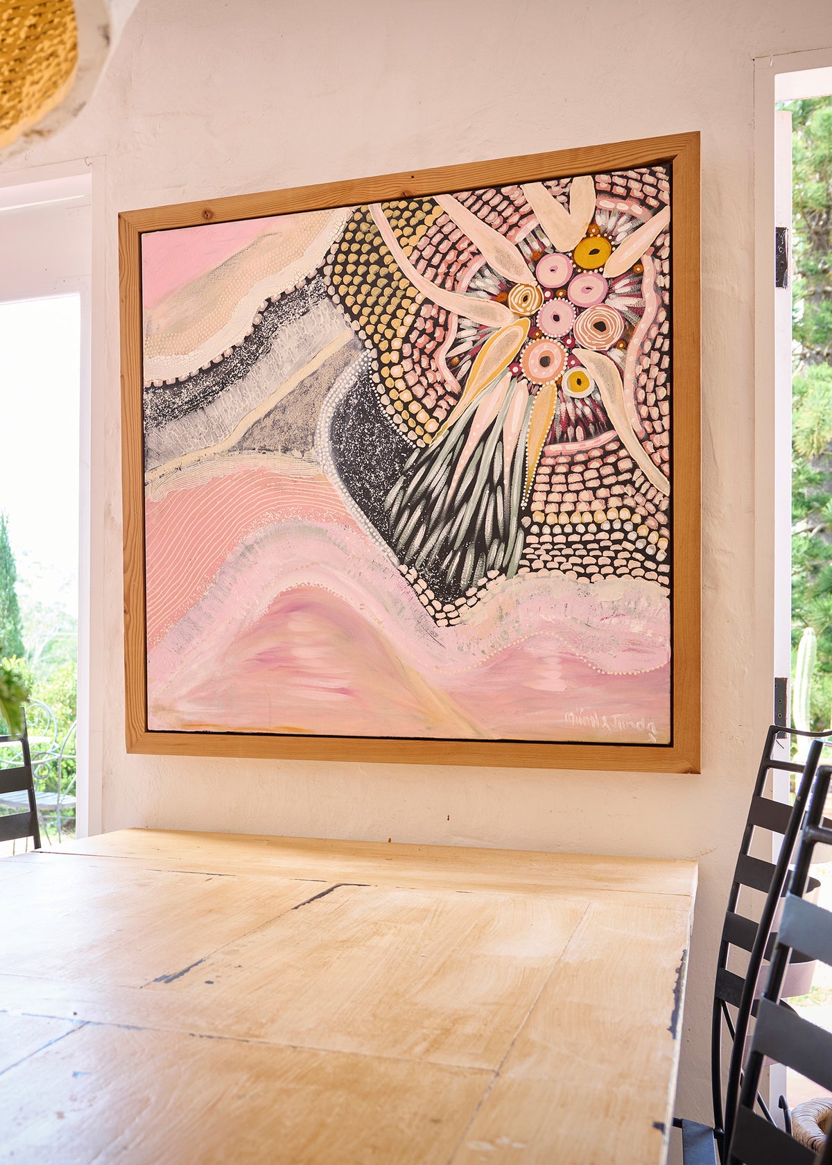 A large framed abstract painting with pink, cream, black, and gold tones hangs on a white wall above a light wood table in a bright indoor setting.