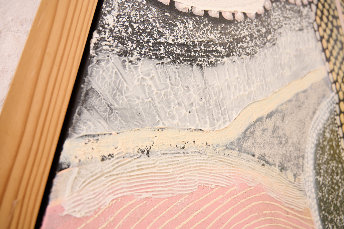Close-up of a textured abstract artwork with layered brushstrokes in gray, cream, pink, and black, framed by a light wood border.