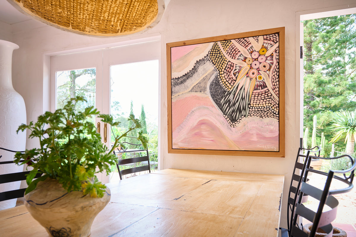 A framed abstract painting with pink, black, and cream patterns hangs on a white wall in a bright dining room with a wooden table, chairs, and a potted plant in the foreground.