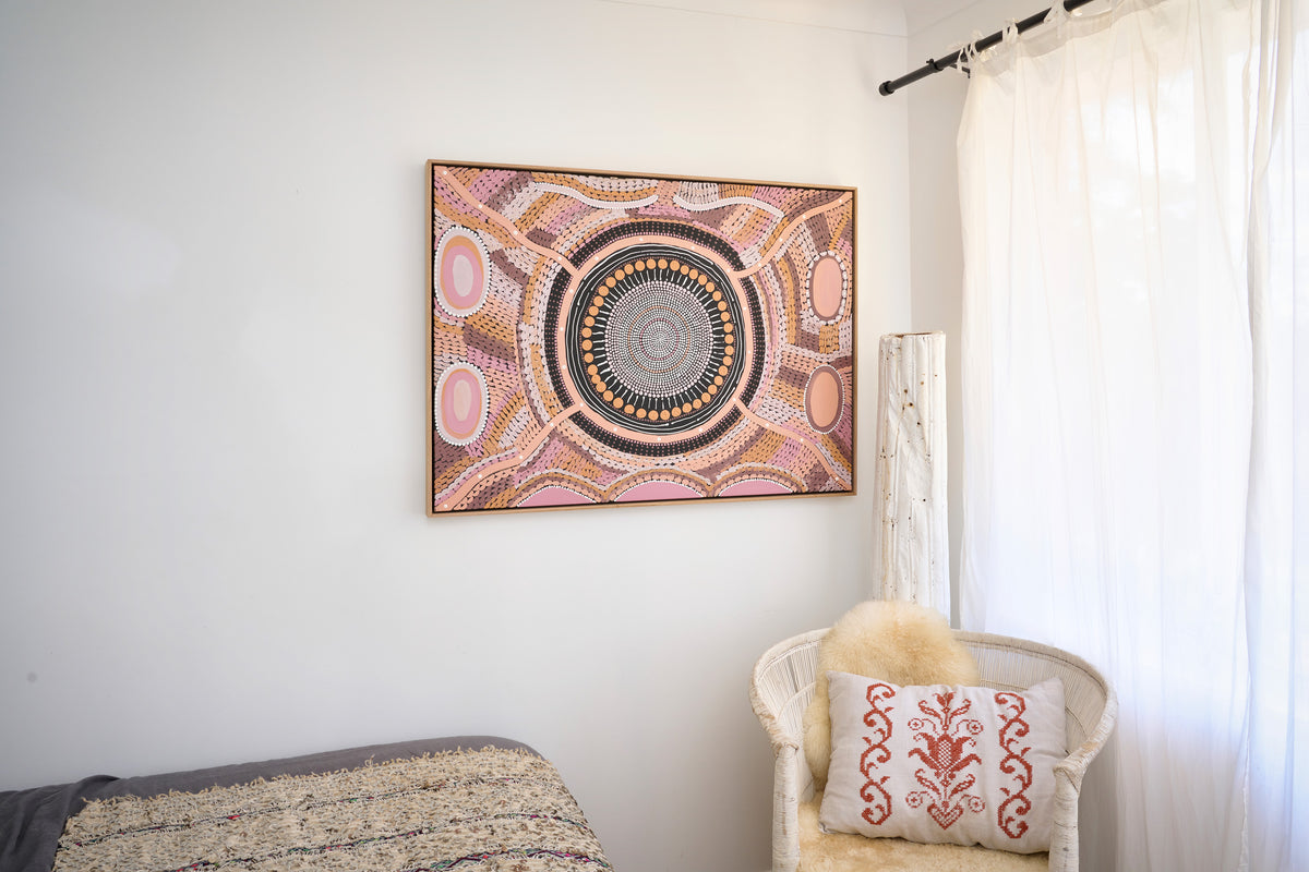 A framed square wall art piece with a circular Aboriginal-style dot pattern in pink, gold, brown, and gray, hanging on a white wall in a bedroom next to a white armchair with a decorative pillow.
