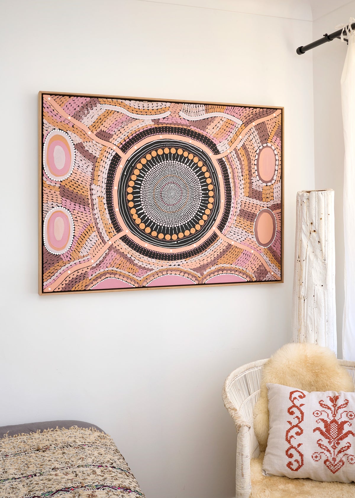 A framed square wall art piece featuring a detailed circular Aboriginal-style dot painting in pink, beige, brown, black, and white, hanging on a white wall in a styled room with a chair and pillow.