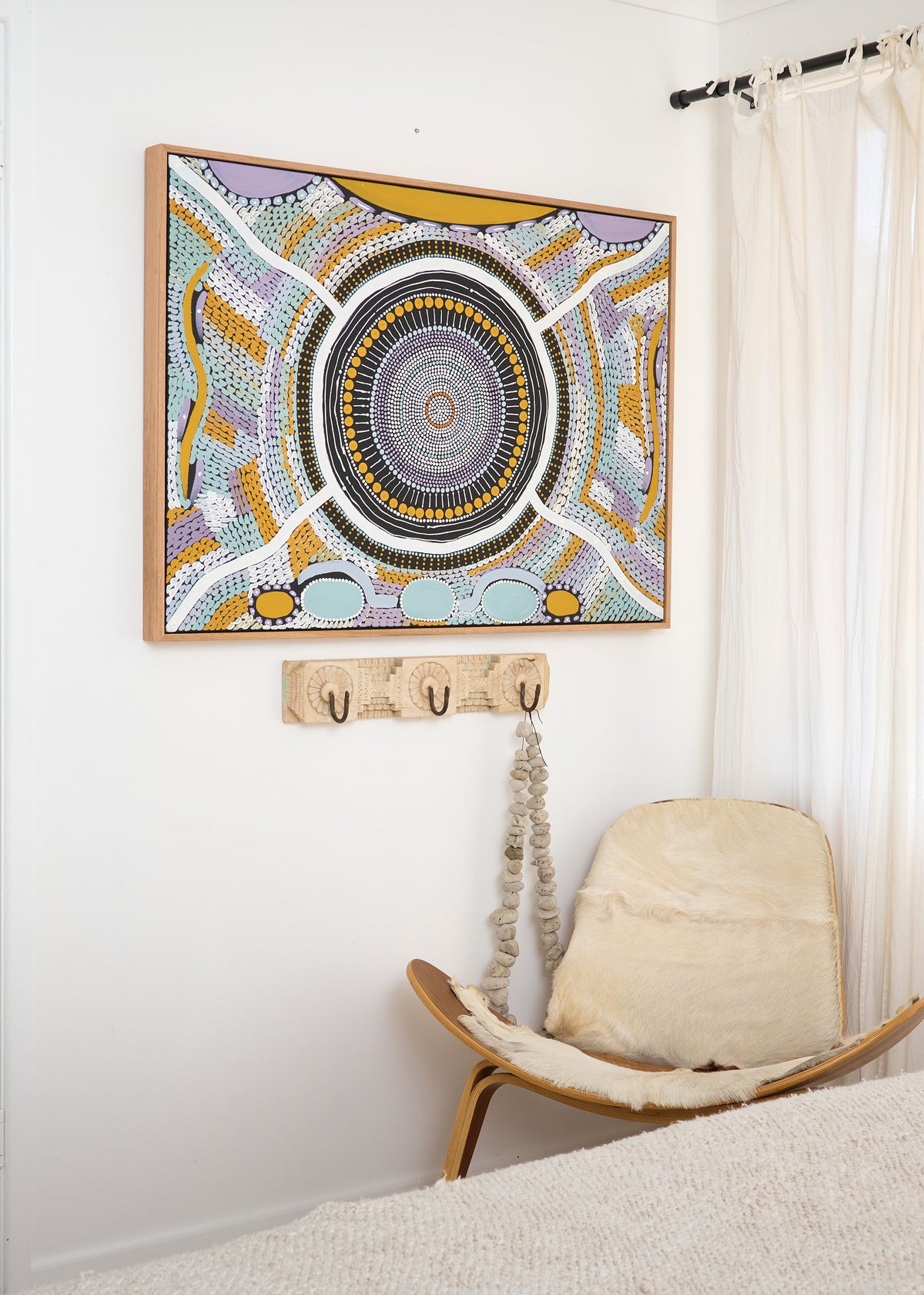 A framed abstract artwork with concentric circular patterns in yellow, gray, purple, black, and white, hanging on a white wall above a wooden coat rack and a cream-colored chair in a bright room.