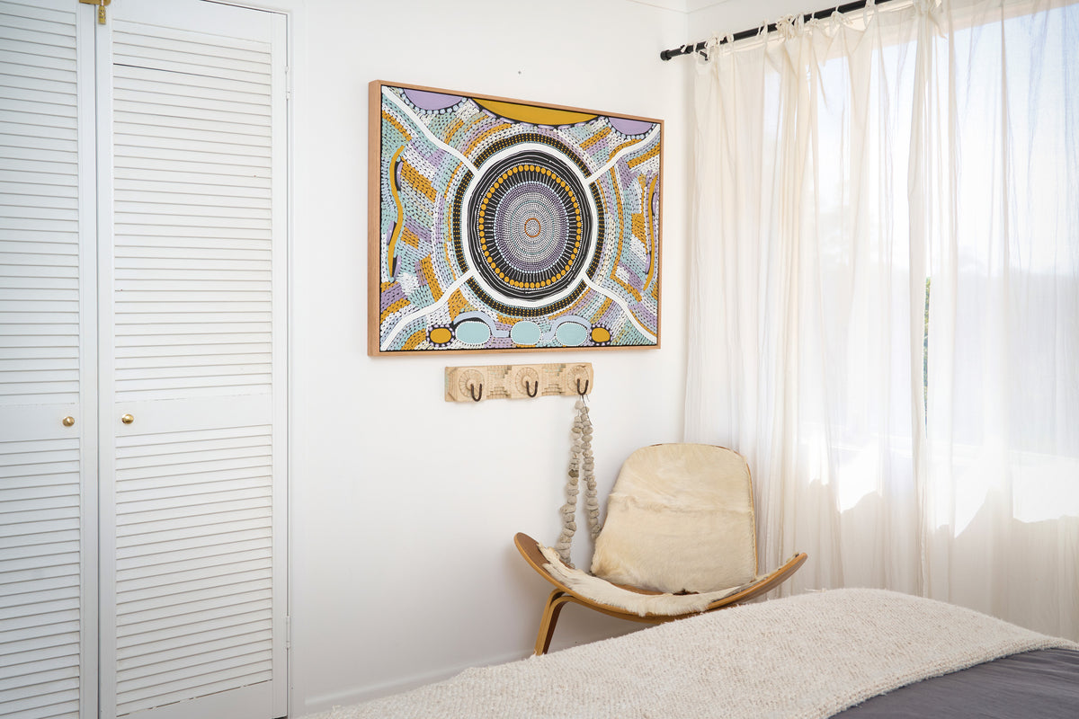 A framed square wall art piece with a circular mandala-style design in blue, yellow, white, and gray, hanging on a white wall in a bedroom above a wooden chair and next to sheer curtains.