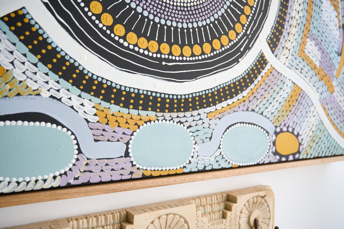 A close-up of a colorful circular Aboriginal-style wall art piece with concentric patterns in black, white, yellow, blue, and lavender, mounted on a light wood frame above a carved wooden object.