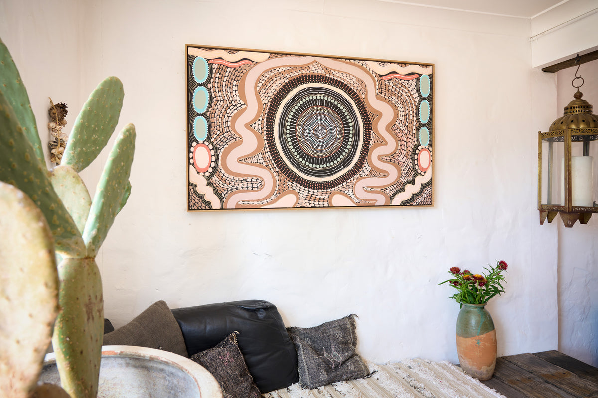 A framed Aboriginal-style wall art piece with intricate circular dot art and wavy lines in beige, brown, black, and muted teal, hanging on a white wall above a sofa in a styled living room.