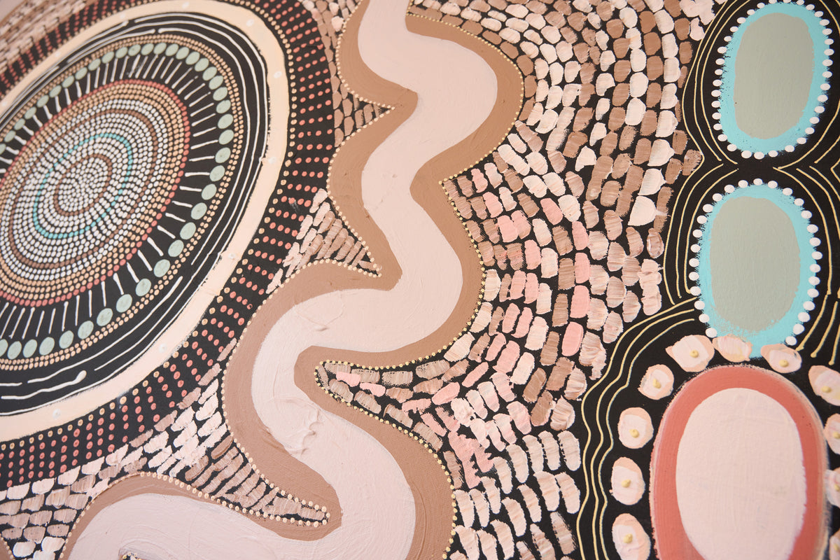 A close-up of a textured Aboriginal-style wall painting or mural featuring a winding pale pink river, concentric dot circles, and organic oval shapes in black, beige, pink, brown, light blue, and white.