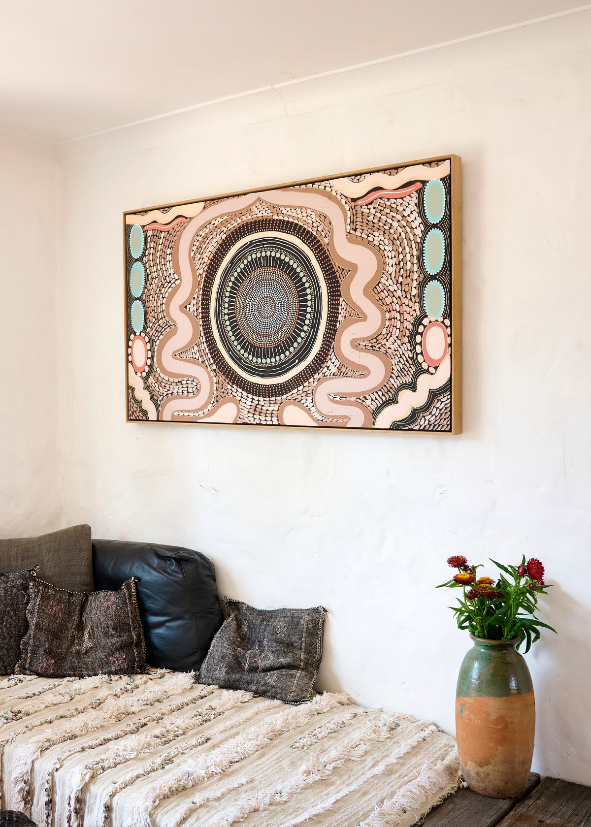 A framed Aboriginal-style circular dot art print hangs on a white wall above a bed with textured pillows and a throw blanket, next to a vase of flowers.