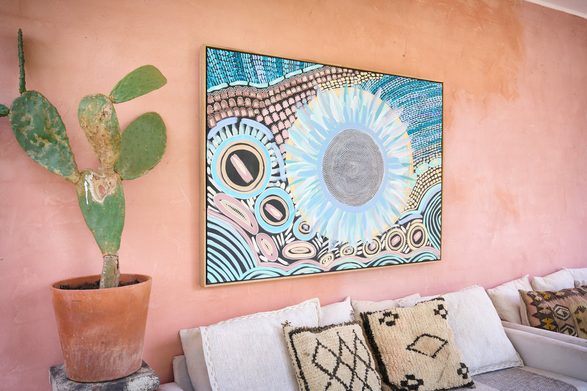 A framed square wall art print with a colorful abstract sun and circular pattern hangs on a peach-pink stucco wall above a white sofa with patterned pillows. A potted cactus stands to the left.