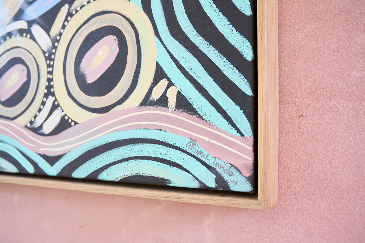 A close-up of a framed abstract painting featuring black, light blue, cream, and pink curved patterns, mounted on a dusty pink wall.