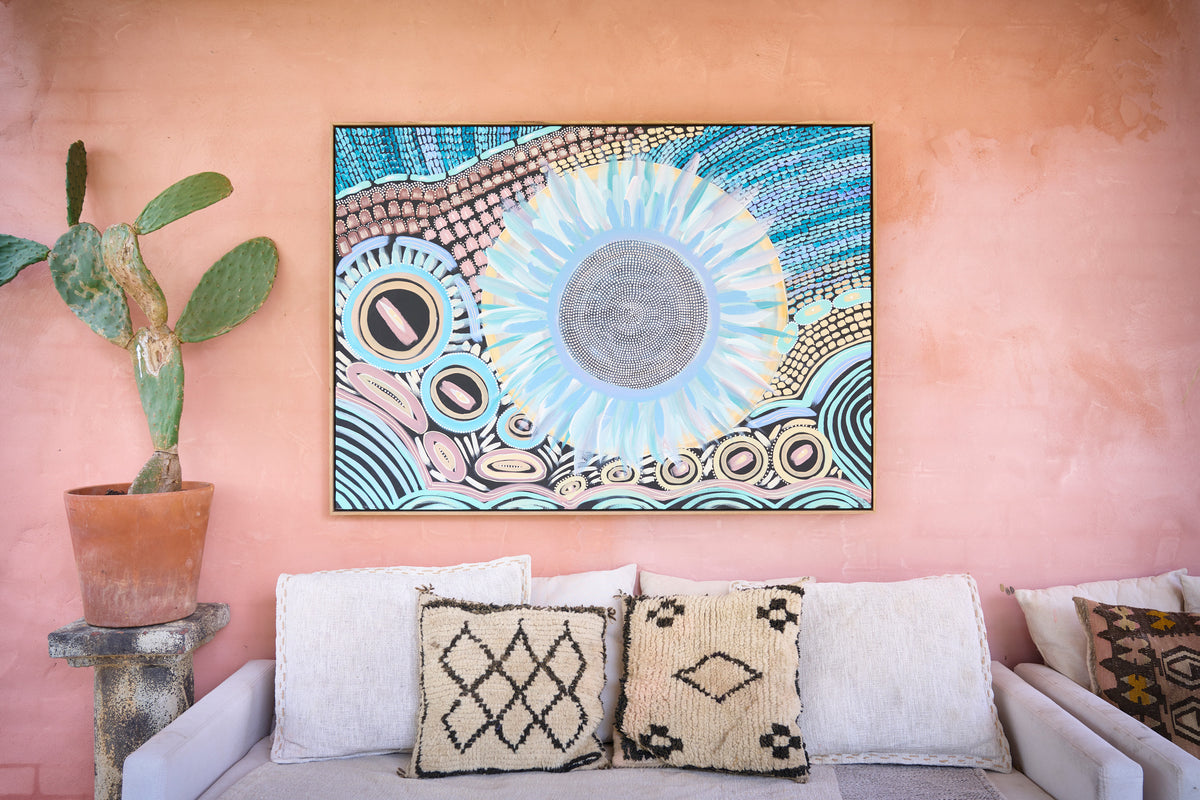 A square framed art print featuring a colorful sun and geometric patterns, displayed on a textured peach-pink wall above a white sofa with patterned pillows and a potted cactus.