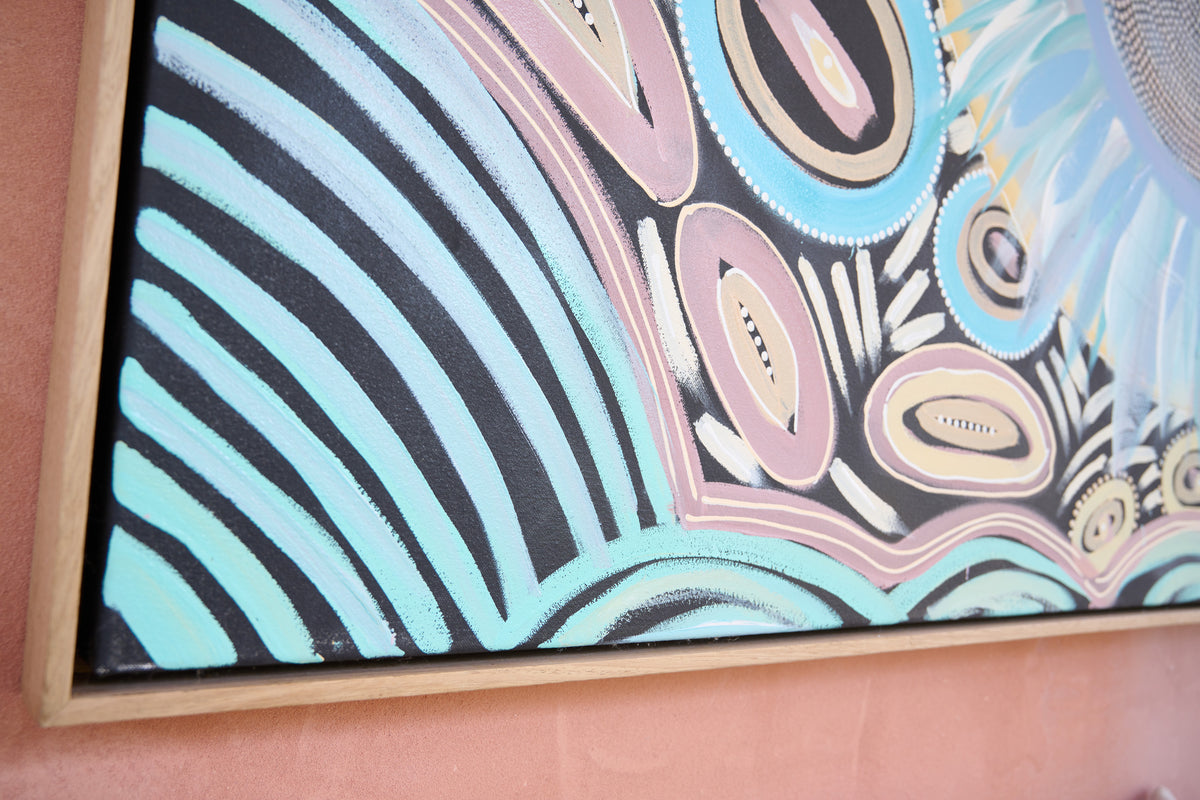 A close-up of a framed abstract painting mounted on a peach-pink wall. The artwork features bold black and light blue curved stripes on the left, with pink, beige, and light blue oval and circular patterns on the right, enclosed in a light natural wood frame.