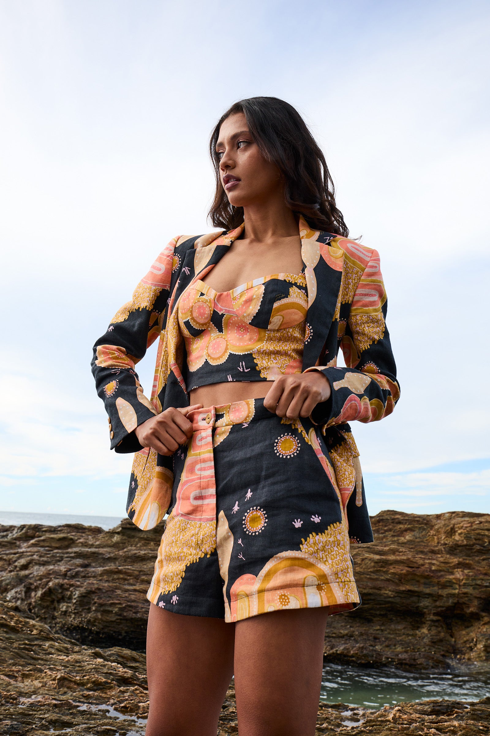 A woman standing on rocky beach wearing a floral print linen slimline comfort jacket and shorts.
